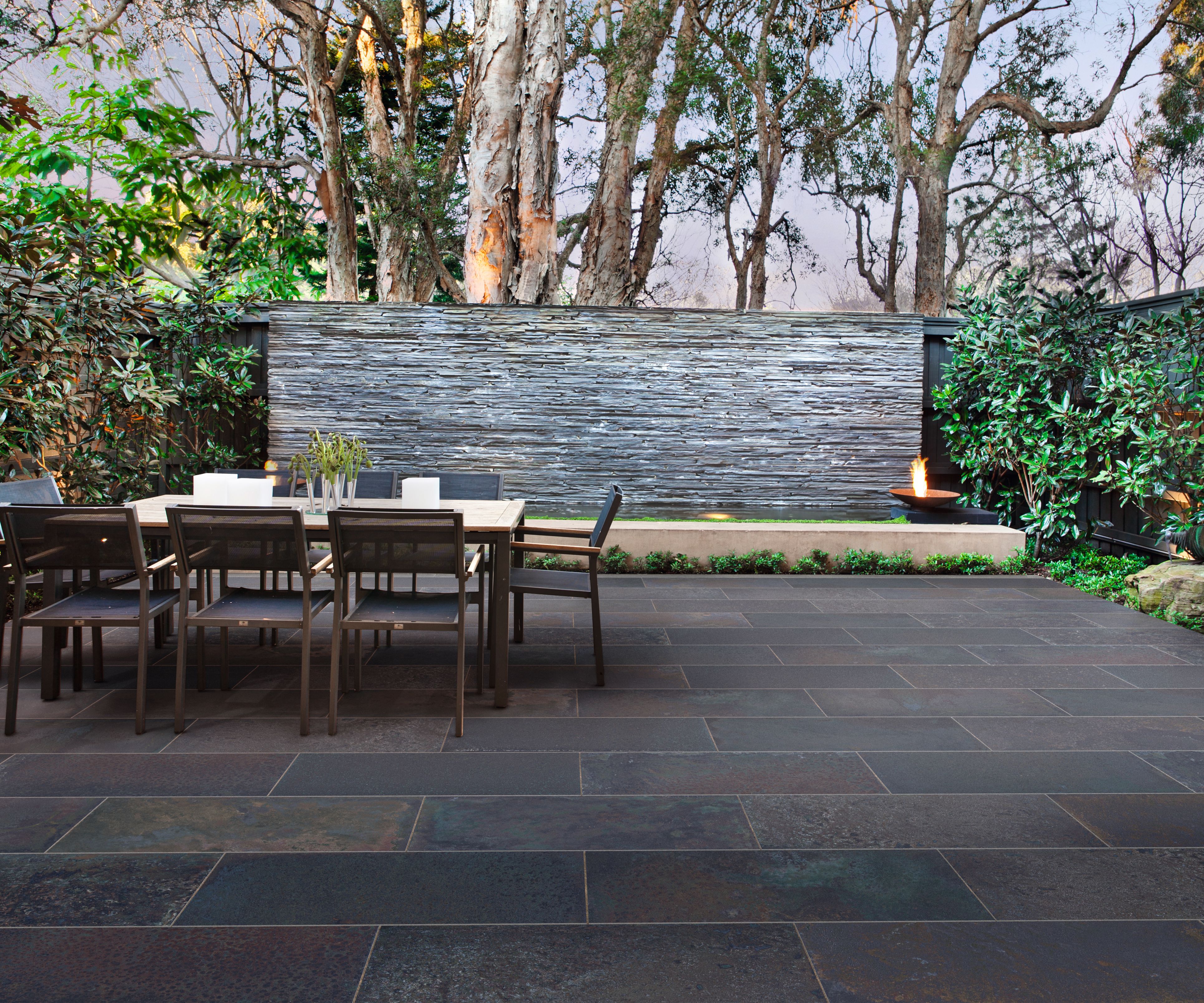SYMPHONY® Elements Corten in Cobalt A dark-tiled patio with a table and chairs in front of a water feature